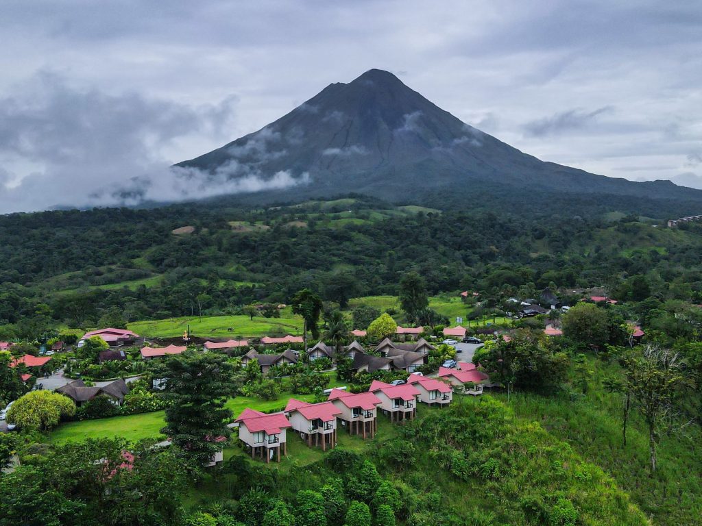 A scenic village with vibrant rooftops nestled below a towering volcano under a cloudy sky.